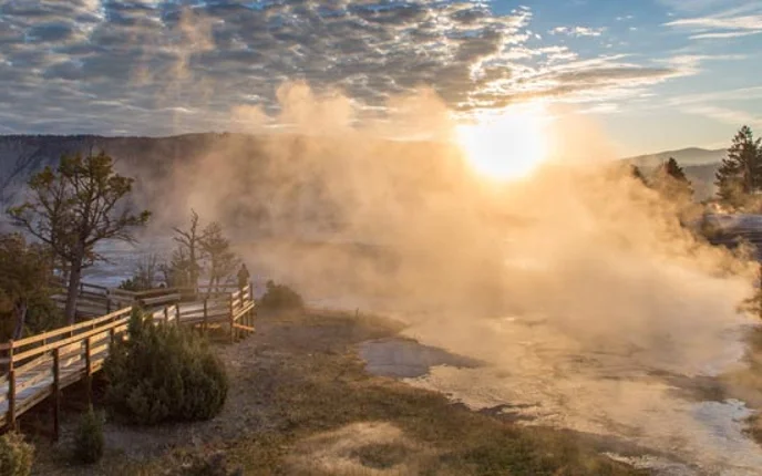 A visitor standing on a boardwalk watches the sunrise through mist rising from the Mammoth Hot Springs.